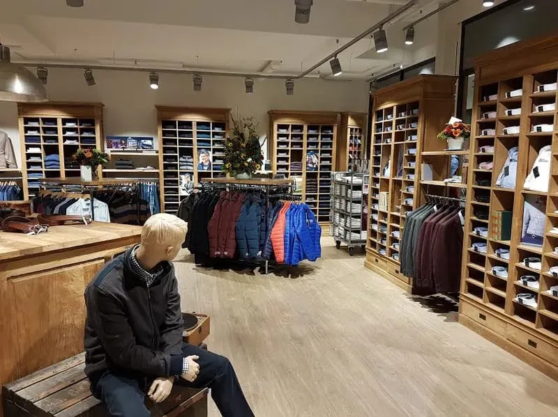 Interieur van de winkel: houten wandkasten met gevouwen overhemden, rekken met jassen en een etalagepop op een oude reiskist.