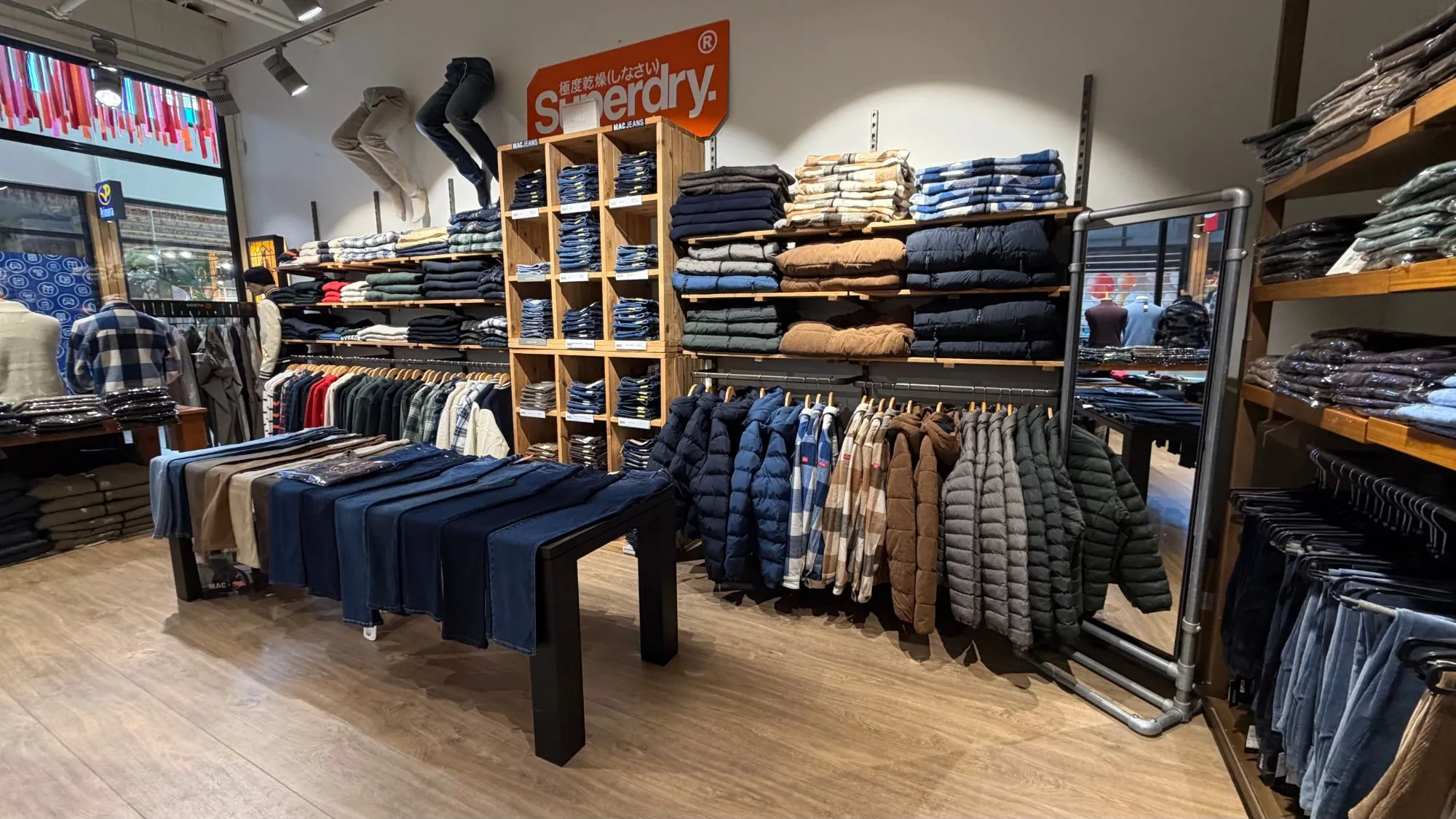Wand met stapels jeans, geruite overhemden en gewatteerde jassen onder het oranje Superdry-logo, met een tafel vol jeans ervoor.