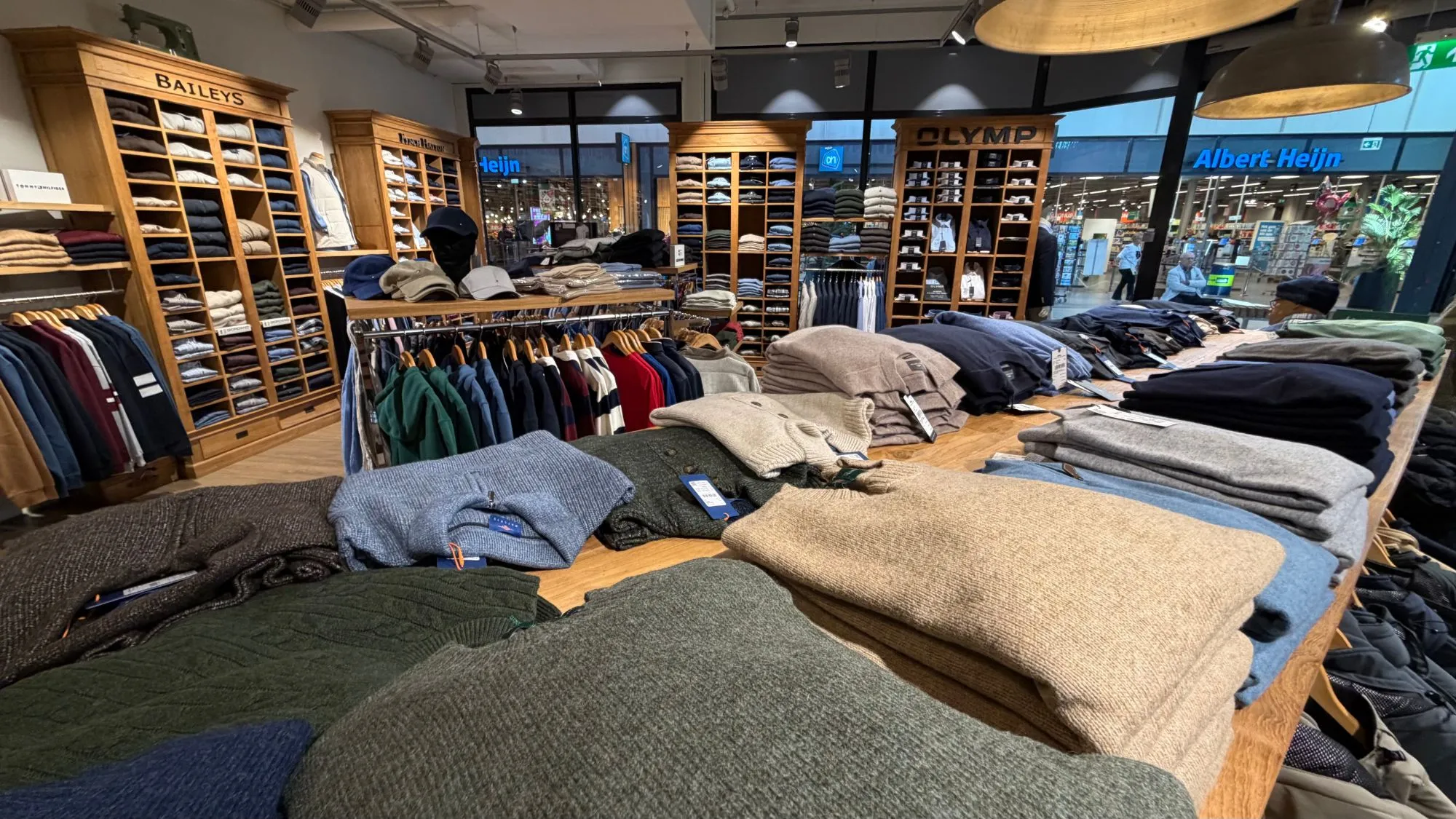 Een lange houten tafel met stapels gebreide truien in groen, blauw en beige, met daarachter de kasten van Baileys en OLYMP.