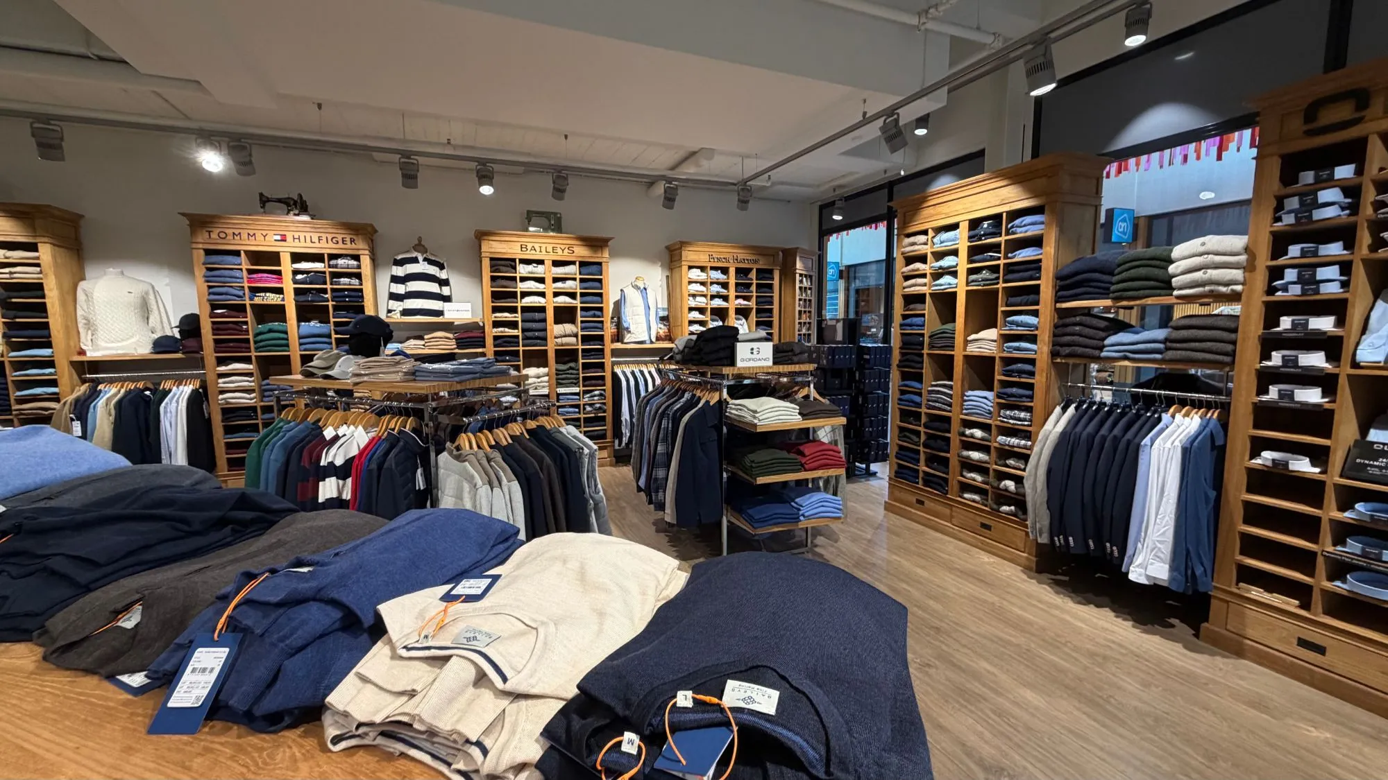 Overzicht van de winkel: houten wandkasten van Tommy Hilfiger, Baileys en Fynch-Hatton vol gevouwen truien en overhemden, met rekken jassen en colberts ervoor.